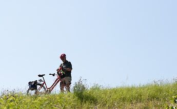 A person standing on a grassy hill inspecting a bicycle, with a clear blue sky above.
