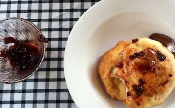 A pastry drizzled with jam on a white plate next to a glass container of more jam on a checkered…