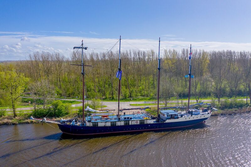 A large sailing vessel named Leafde fan Fryslân is docked alongside a scenic tree-lined canal, under a bright blue sky. (AI Description)