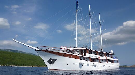 A large white sailing vessel named "Princeza Diana" is on the water against a backdrop of green hills and a blue sky.