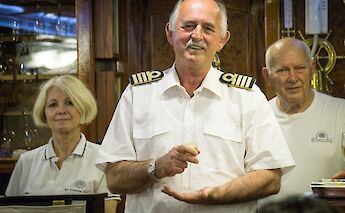 A person wearing a captain's uniform stands smiling, holding a glass, with two others nearby…