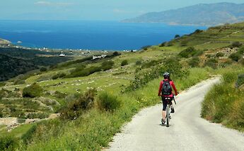 A cyclist in a red jacket rides along a winding road with lush green hills and a sea view under a…