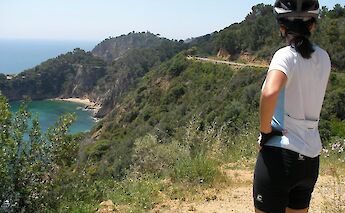 A person in cycling gear stands observing a coastal landscape with a cliff, greenery, and a beach…