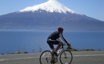 A cyclist rides along a road with a snow-capped mountain and a body of water in the background.