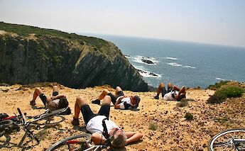 People rest beside their bicycles on a cliff overlooking a rocky coastline and ocean.
