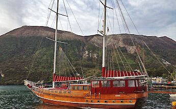 A wooden sailing boat named Sundial docked by the shore with mountainous terrain in the background.