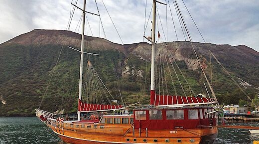 A wooden sailing boat named Sundial docked by the shore with mountainous terrain in the background.