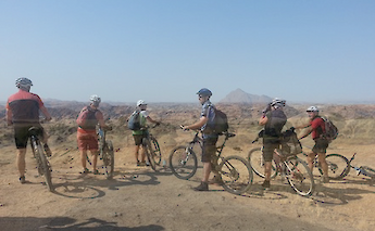A group of cyclists standing on rocky terrain, looking towards distant hills under a clear sky.