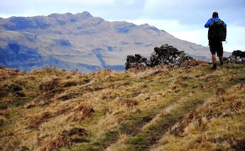 A person wearing a backpack walking on a grassy hillside with distant mountains under a cloudy sky.