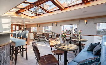 The lounge area on the Merlijn, featuring chairs, tables, and large windows.