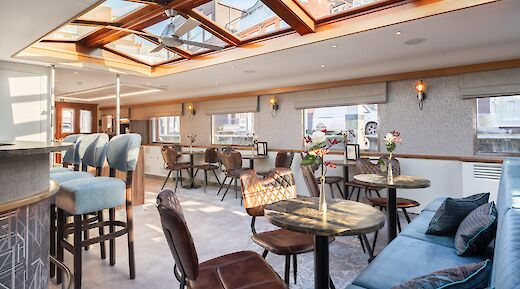 The lounge area on the Merlijn, featuring chairs, tables, and large windows.