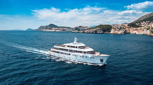 A boat named Yolo sailing in clear blue waters near the Croatian coastline, with picturesque cliffs and a small town visible in the distance.
