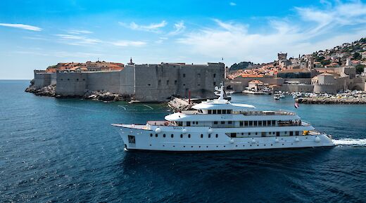 A large white boat sails near the historic city walls and structures of Dubrovnik, Croatia, under a clear blue sky.