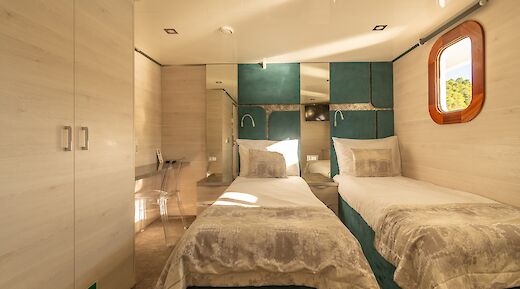 A cozy twin cabin on a boat with modern furnishings, featuring two single beds, a window with a view, and a desk with a transparent chair.