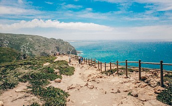 A rocky coastal path at Cabo da Roca, Portugal, with visitors walking along the fenced trail above the ocean.