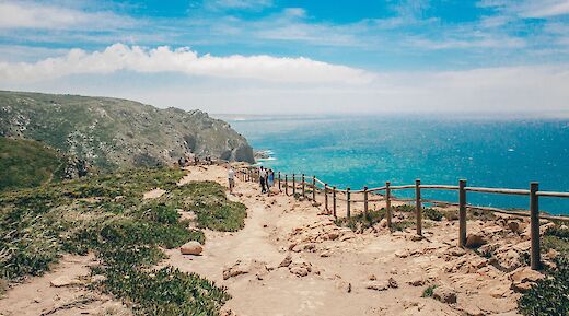 A rocky coastal path at Cabo da Roca, Portugal, with visitors walking along the fenced trail above the ocean.