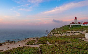 A lighthouse stands on a cliff overlooking the ocean, with a vibrant sunset sky and greenery on the landscape.