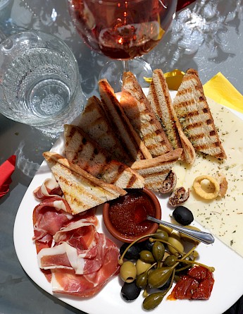 A plate featuring grilled bread, cured meats, olive, and a small dish of sauce, viewed from above.