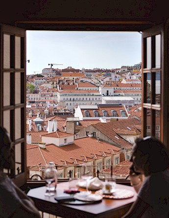 A view of Lisbon's rooftop with red tiles seen through a window, with a table in the foreground.