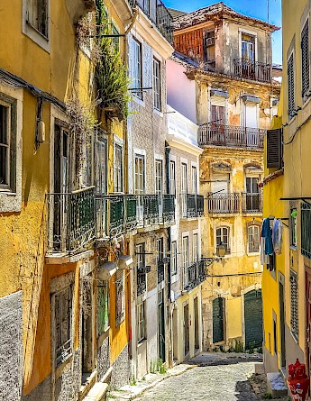 A narrow, cobblestone street in Lisbon is lined with colorful, weathered buildings featuring balconies and greenery.