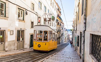A yellow tram traveling up a steep, narrow street in Lisbon, Portugal, surrounded by traditional buildings.