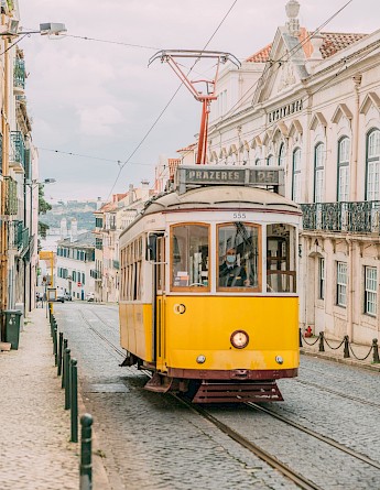A yellow tram travels down a cobblestone street lined with pastel-colored buildings in Lisbon, Portugal.
