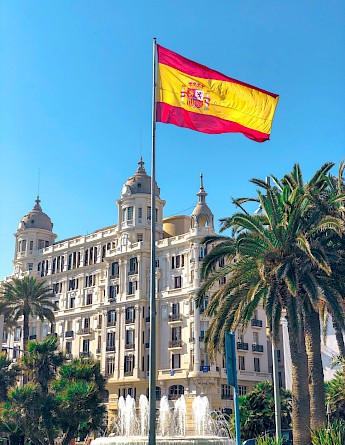 A palm-lined plaza in Madrid features a large Spanish flag flying in front of an ornate, historic building and a fountain.