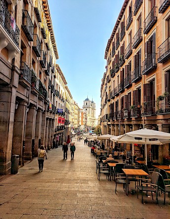 A sunlit street scene in Madrid, Spain, lined with historic buildings and outdoor cafes.