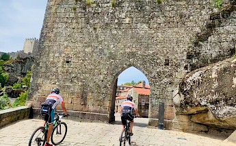 Two cyclists approach a large stone archway, with a historic wall and buildings visible beyond.
