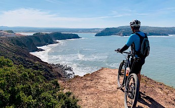 A cyclist with a mountain bike stands on a rocky cliff overlooking the ocean, with rugged coastline visible in the distance.