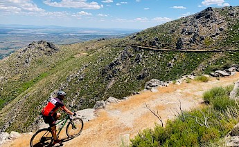 A cyclist rides along a rugged mountain trail with expansive views of surrounding hills and a clear sky.