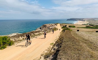 Cyclists ride along a coastal dirt path with the ocean to the left and rolling hills on the right.