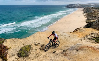 A cyclist on a dirt trail along a cliff, overlooking a beach and the ocean.
