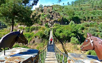 A suspension bridge with decorative horse statues leads to a hillside with stone ruins and lush greenery. A person is walking a bike across the bridge.