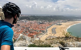 A cyclist wearing a helmet and blue jersey overlooks the coastal town of Nazaré, Portugal, with a view of its sandy beach and ocean.
