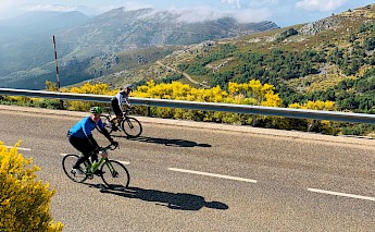 Two cyclists ride along a mountain road with scenic views of hills and forests under a clear sky.