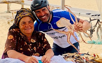 A smiling cyclist wearing a helmet holding a dried octopus, standing next to a woman sitting at a table on a sandy beach.