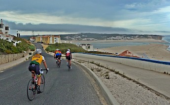 Cyclists ride down a road along a coastal area with a beach and buildings visible in the background.