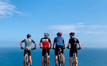 Four cyclists wearing helmets and gear stand facing the ocean, with their bikes beside them.