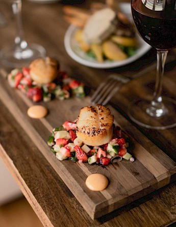 A dining setup featuring a wooden board with seared scallops on a bed of chopped vegetables, accompanied by glasses of red and white wine.