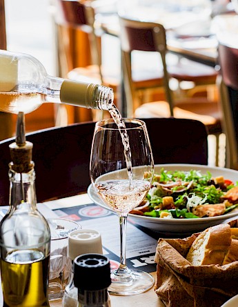 A bottle of white wine being poured into a glass, with a salad and bread on a table.
