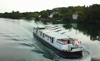 A boat named Clair de Lune is traveling on a river, with trees and houses lining the banks.