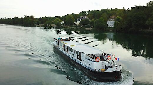 A boat named Clair de Lune is traveling on a river, with trees and houses lining the banks.