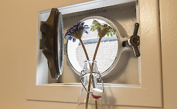 A round porthole window with metal fixtures, featuring two flowers in a glass bottle in front of it.