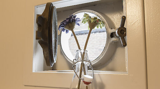 A round porthole window with metal fixtures, featuring two flowers in a glass bottle in front of it.
