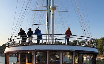 Four people standing on the deck of a boat with tall masts, leaning on the railing and gazing out at the water, amidst a backdrop of clear skies and a wooded shoreline.