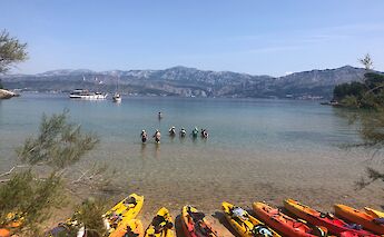A serene beach scene with colorful kayaks lined up on the sandy shore, people wading in the shallow, clear blue water, and a mountainous landscape in the distance.