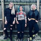 Three individuals stand against a wooden fence, dressed in casual, stylish clothing; two with dyed hair and various fashion accessories.