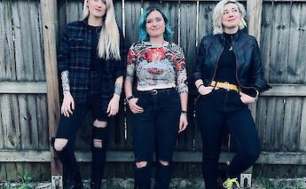 Three individuals stand against a wooden fence, dressed in casual, stylish clothing; two with dyed hair and various fashion accessories.