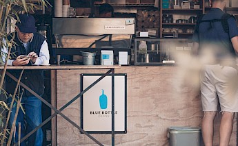 A person using a phone while seated at a cafe counter, with coffee equipment visible in the background.